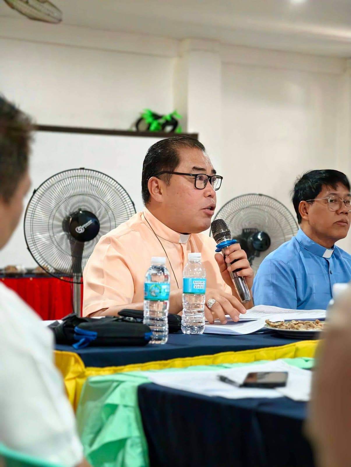 A member of the Board speaking into a microphone at the meeting table, with another trustee seated beside him