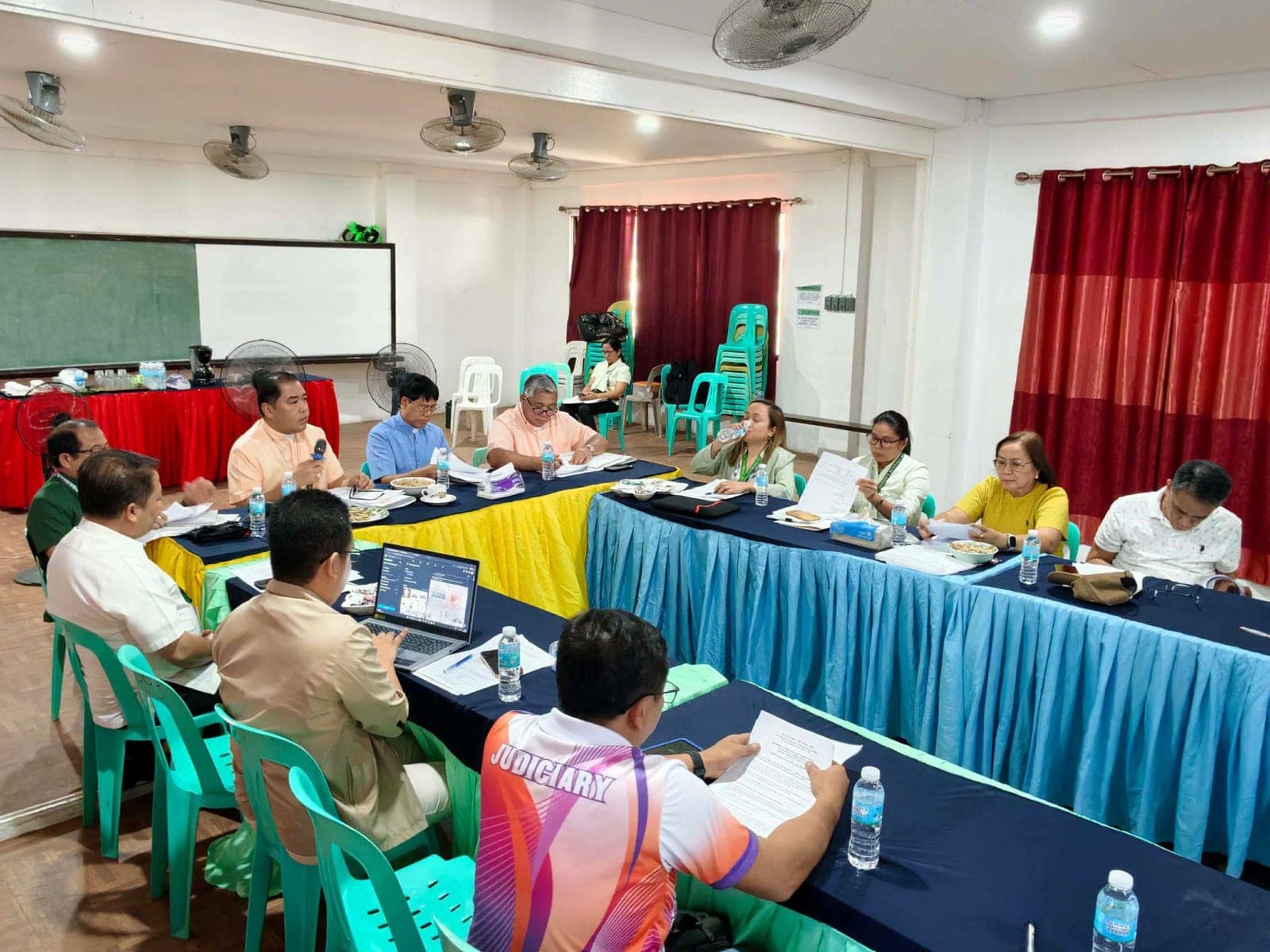 A wide view of the meeting, with trustees seated around draped tables reading documents