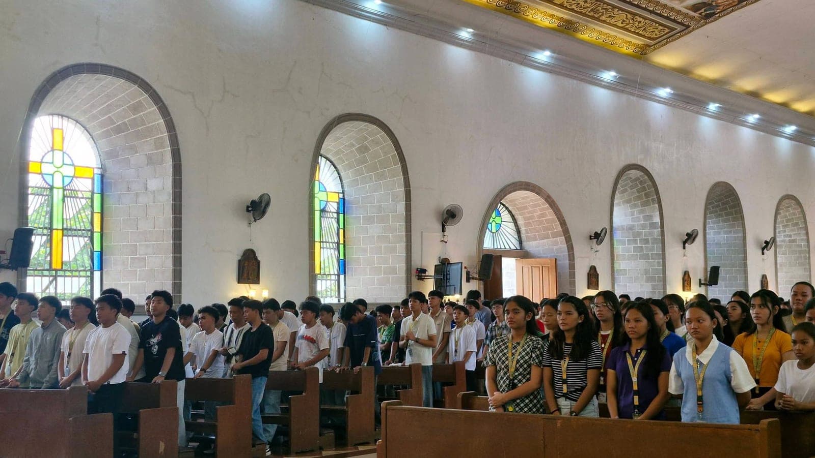 Students stand in prayer in the pews of the parish church as light comes through the stained glass windows
