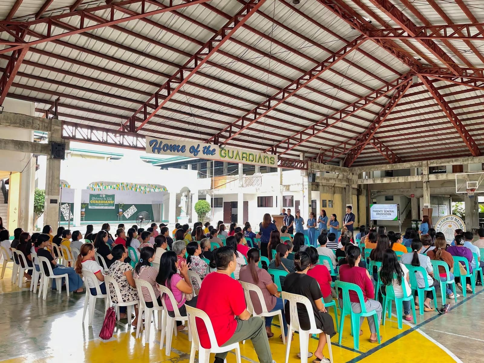 Parents seated in the covered court for the Parents' General Orientation, with teachers standing beneath a banner reading Home of the Guardians