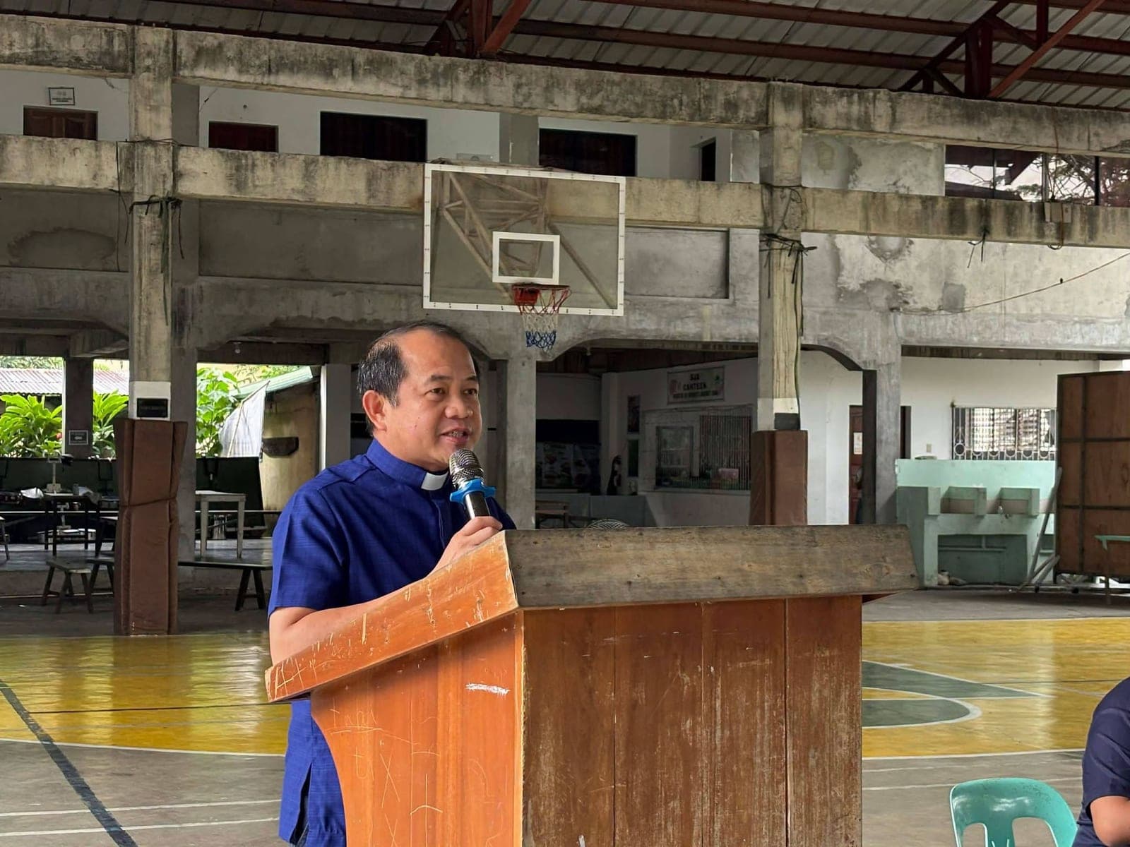 A priest addresses parents from a wooden lectern in the covered court